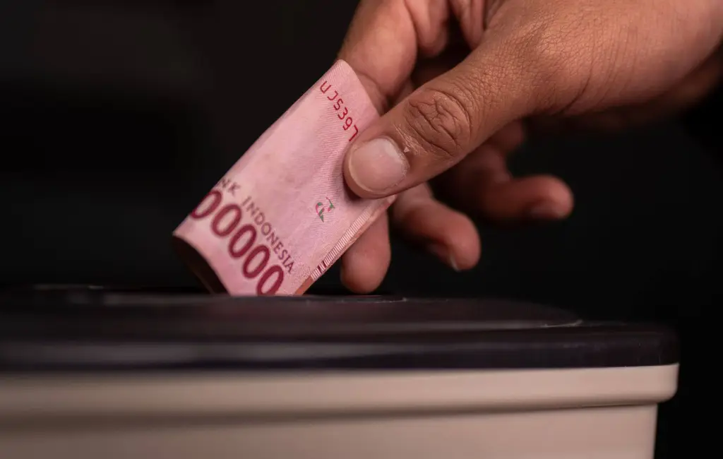 Close-up of a hand donating an Indonesian Rupiah note into a charity box, symbolizing generosity.